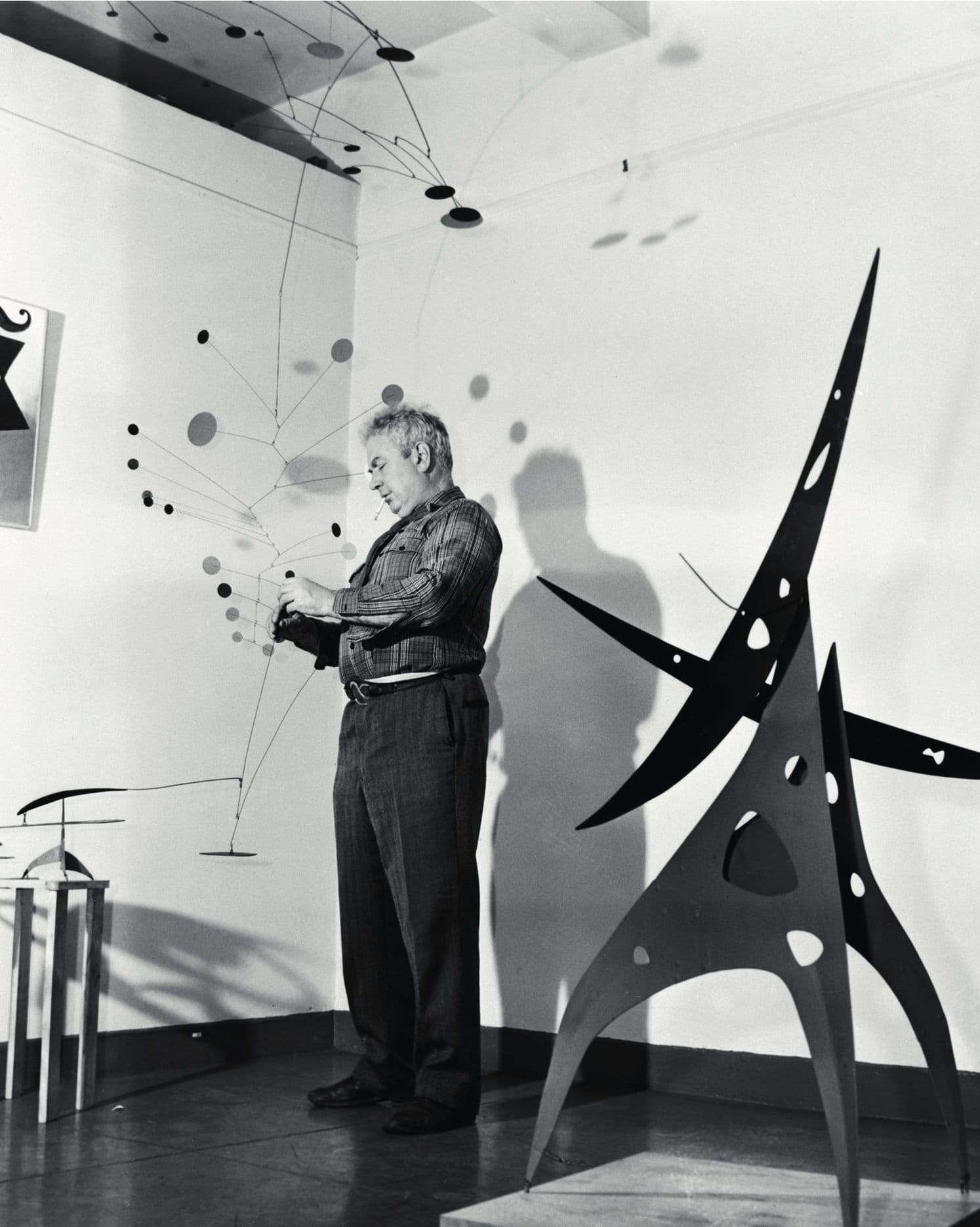 Alexander Calder in his studio, adjusting a hanging mobile whose discs cast shadows across the wall, a large black stabile standing beside him.