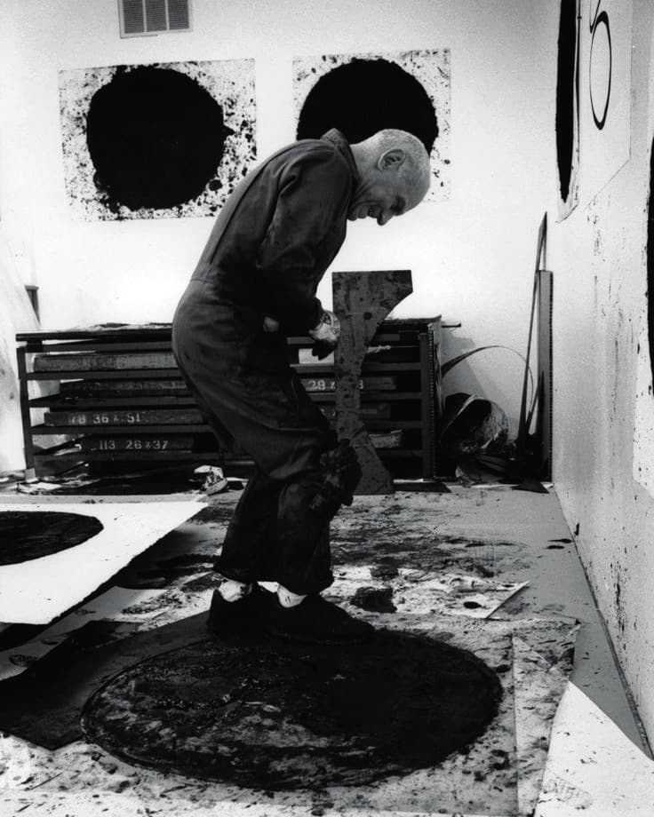 Richard Serra at work in his studio, bent over a large black circular work on the floor, further black works on the wall behind him.