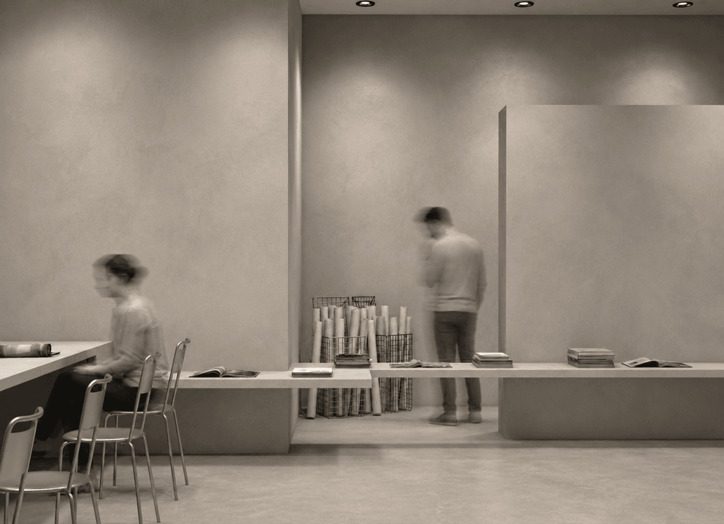 A reading bench with rolled prints in wire baskets, a visitor browsing beside a freestanding concrete panel.